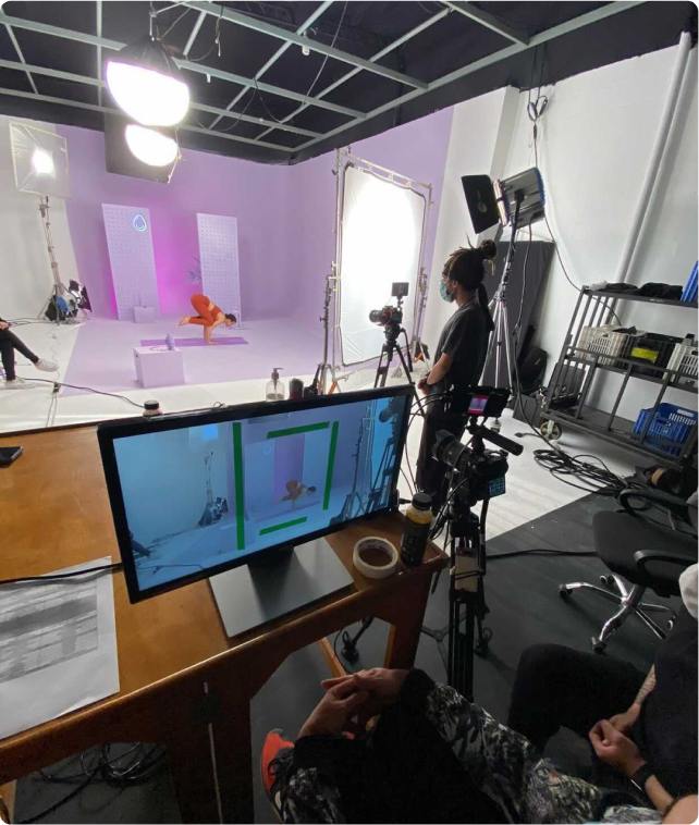 Atriz fazendo posição de Yoga em um estúdio fotográfico, com pessoas atrás de um computador conferindo as fotos